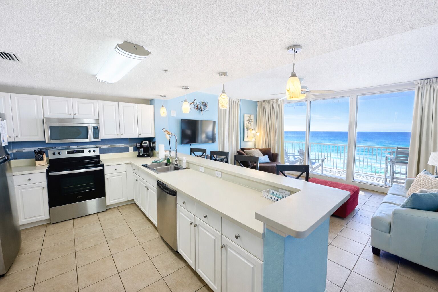 A modern kitchen with white cabinets, a stainless steel oven, and a large island with a sink. There is a dining area with a red table and chairs, and a large window offering a view of the ocean.