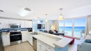 A modern kitchen with white cabinets, a stainless steel oven, and a large island with a sink. There is a dining area with a red table and chairs, and a large window offering a view of the ocean. - Steps to the Sand at our Beachfront Condo at Majestic, 5th floor convenience  (Panama City Beach)