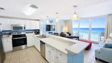 A modern kitchen with white cabinets, a stainless steel oven, and a large island with a sink. There is a dining area with a red table and chairs, and a large window offering a view of the ocean.