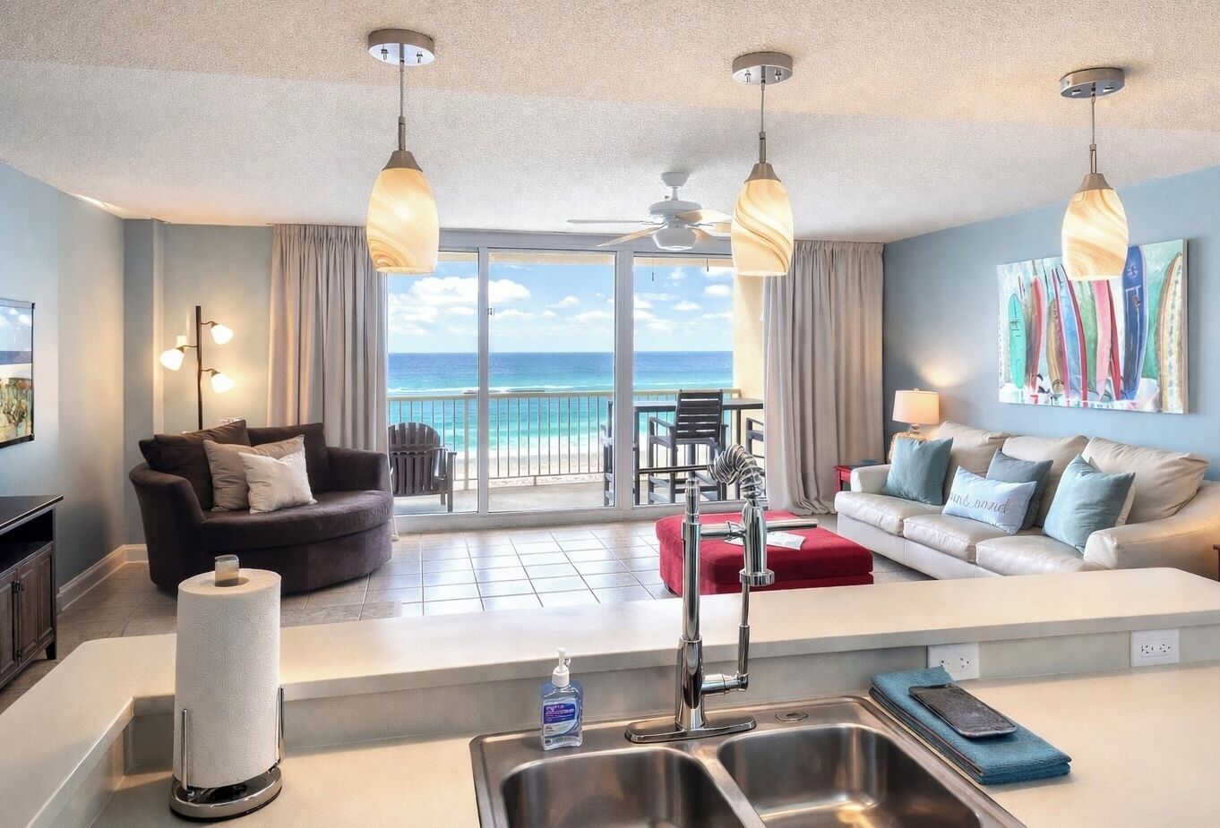 A modern living room with a large window overlooking the ocean, a kitchen area with a sink and a bottle of hand soap, and two sofas facing each other.