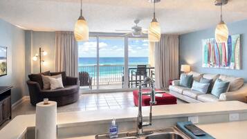 A modern living room with a large window overlooking the ocean, a kitchen area with a sink and a bottle of hand soap, and two sofas facing each other.