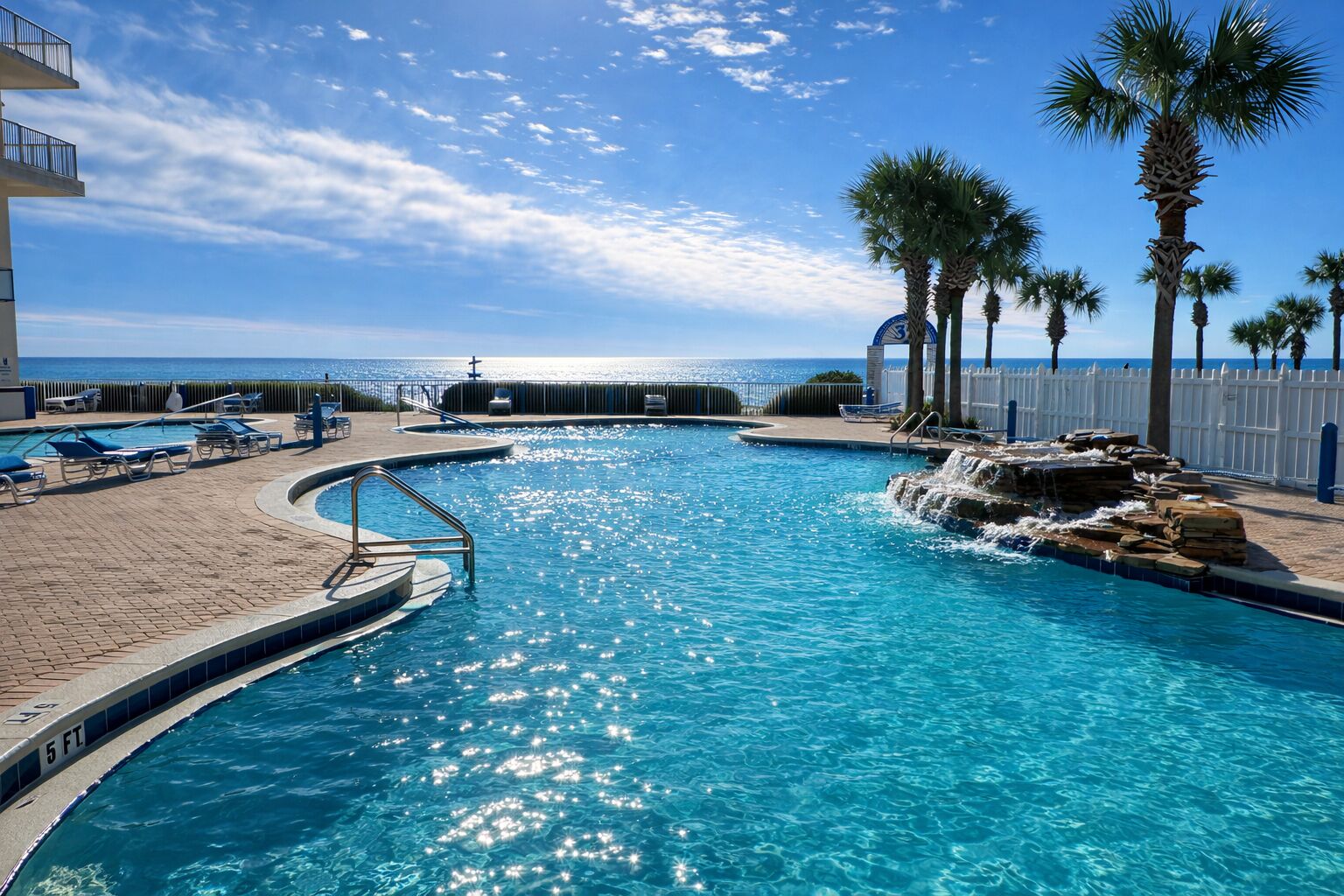 Une piscine dont la profondeur maximale est indiquée à 1,5 mètre, entourée de palmiers et délimitée par une clôture blanche, et offrant une vue sur l’océan.
