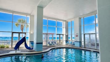 An indoor swimming pool with clear views of the ocean through large windows, a slide, and lounge chairs.