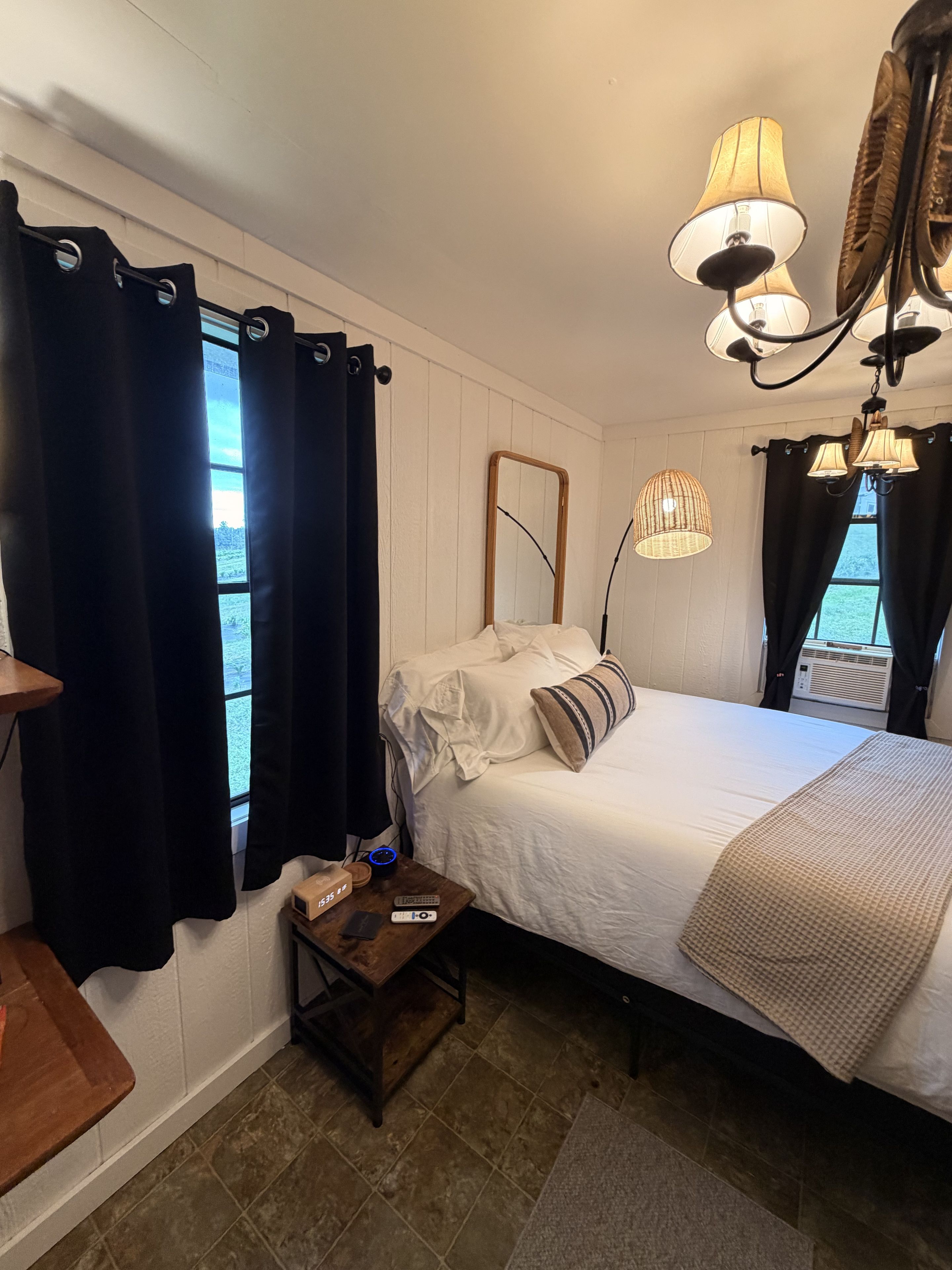 Une chambre à coucher avec un lit, une table de chevet avec une lampe, une fenêtre avec des rideaux et un miroir fixé au mur.