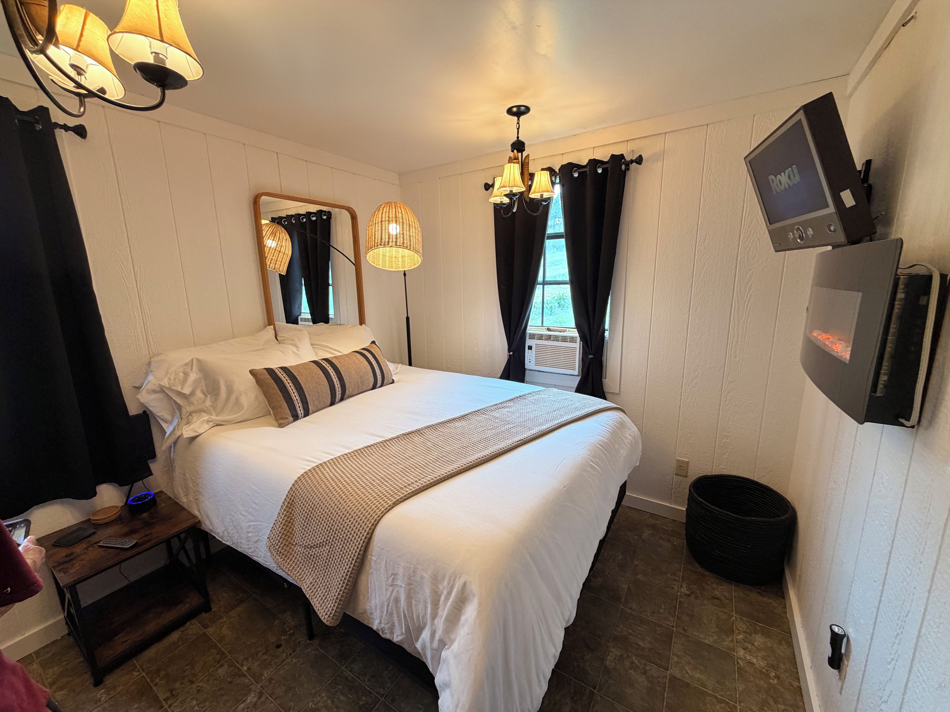 Ein Hotelzimmer mit Bett, Nachttisch, Lampe, Klimaanlage und einem an der Wand montierten Fernseher.