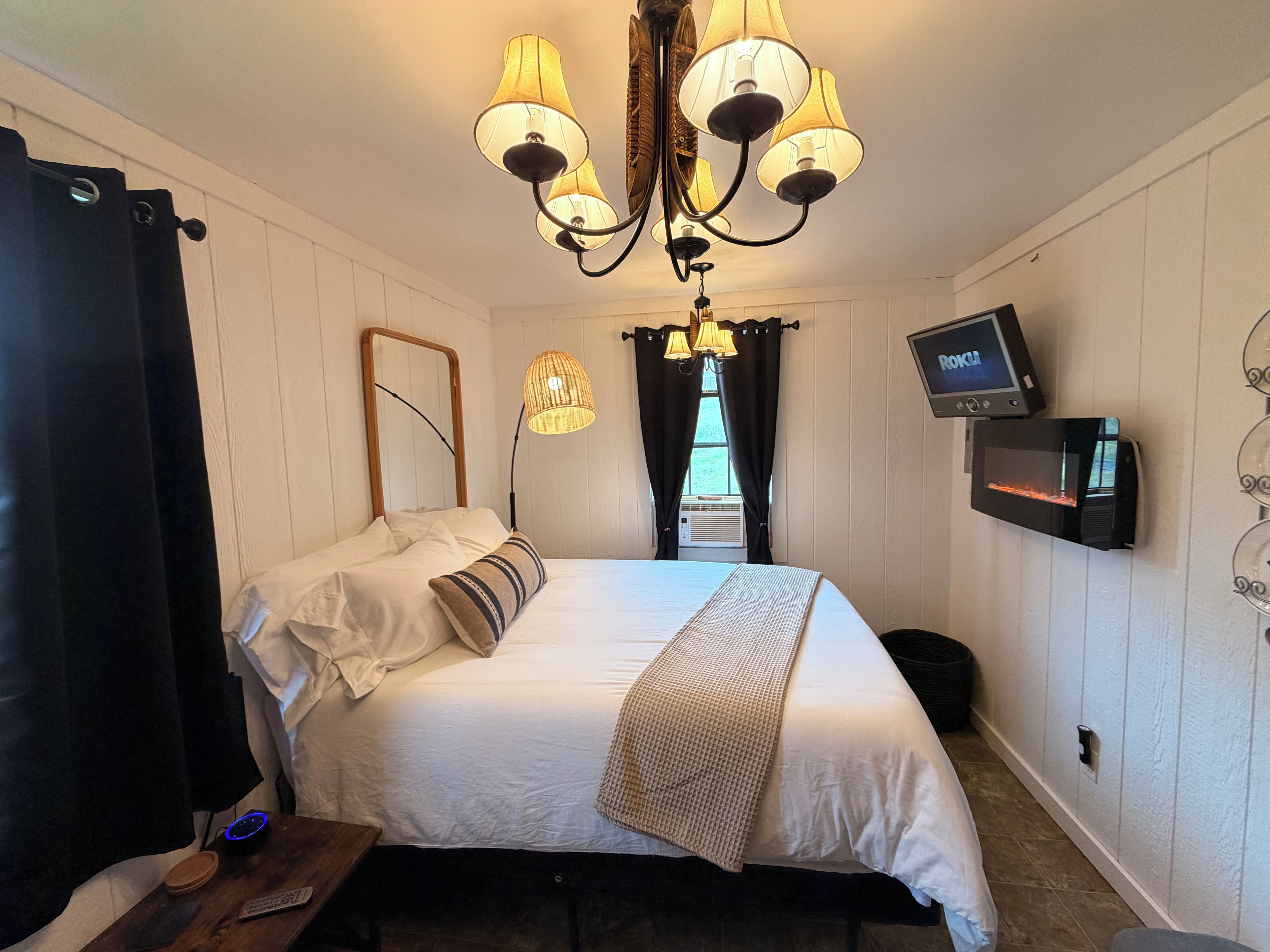 A bedroom with a bed, a television mounted on the wall, a ceiling light fixture, and a window with curtains.