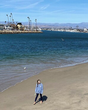 A beach with a person walking on the sand, a rocky breakwater with boats, and mountains in the background. - Charming Silverstrand Beach Cottage Just Steps to the Sand! (Oxnard)