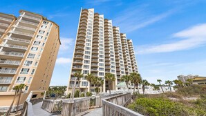 A high-rise residential building with multiple balconies, a wooden boardwalk, and palm trees. - Spectacular Views-Gulf Front Corner unit - Hidden Dunes Gulfside 1506 (Miramar Beach)