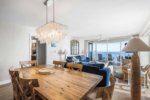 Ein Essbereich mit Holztisch und Stühlen, einer Chandelüster, einer Couch mit Kissen und Blick auf das Meer durch große Fenster.
