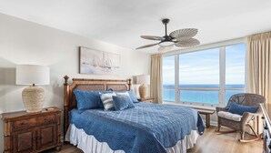 A bedroom with a large bed, a ceiling fan, a view of the sea, a wooden nightstand, and a wicker chair. - Spectacular Views-Gulf Front Corner unit - Hidden Dunes Gulfside 1506 (Miramar Beach)