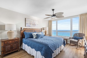 A bedroom with a large bed, a ceiling fan, a view of the sea, a wooden nightstand, and a wicker chair.