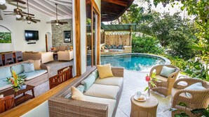 Zona junto a la piscina con mobiliario de mimbre, un televisor y un amplio ventanal con vistas a la piscina.
