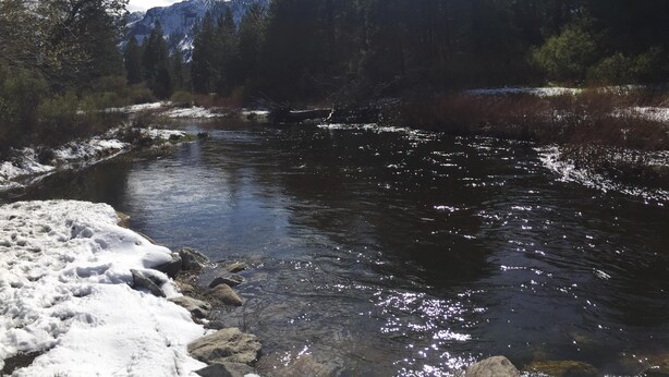Un río con agua cristalina que refleja la luz del sol, rodeado de terreno cubierto de nieve y un bosque denso.