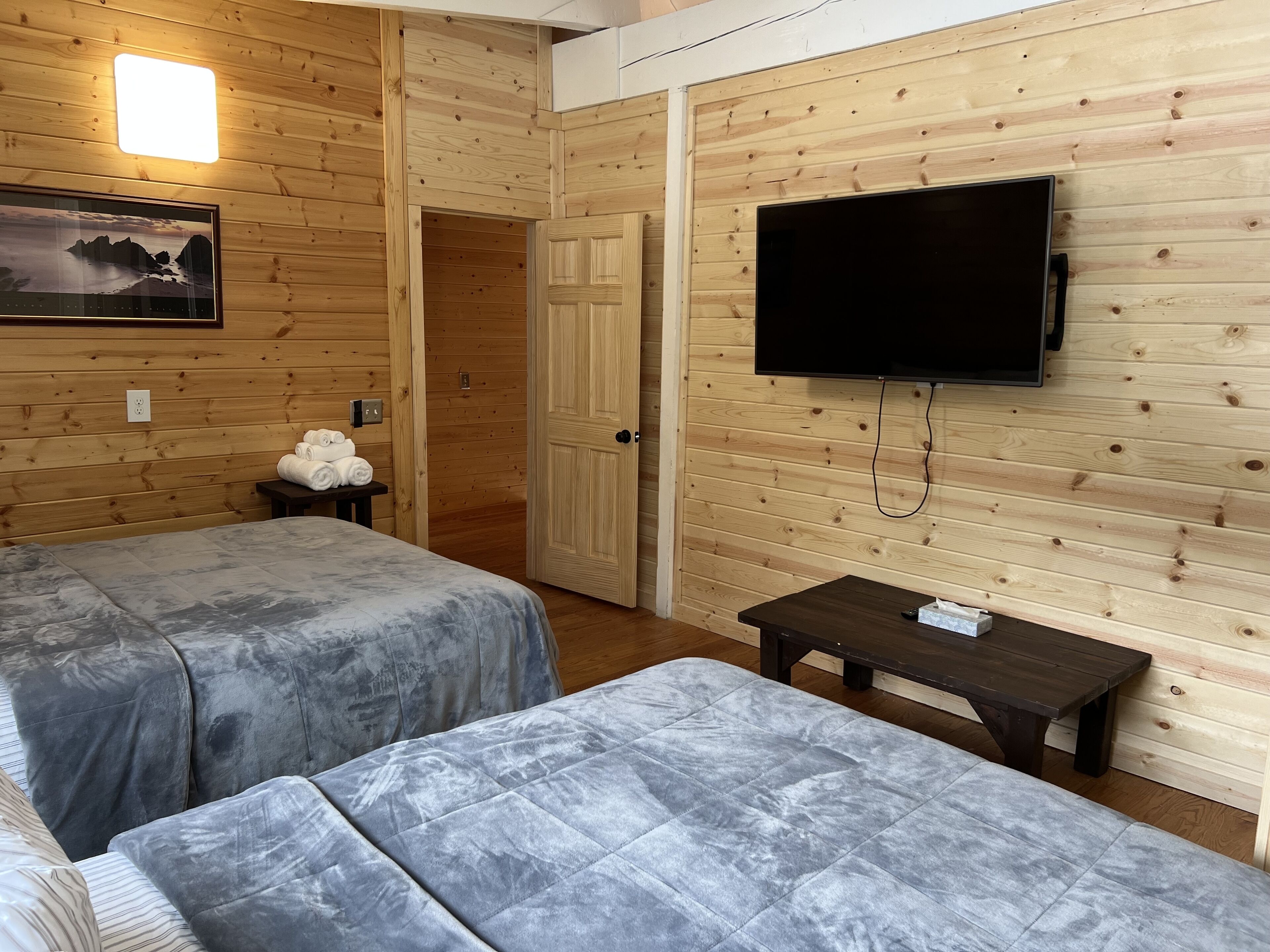 A room with wooden panel walls, two beds with grey covers, a flat-screen TV mounted on the wall, and a small table with a remote control.