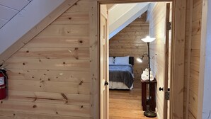 Un interior de madera con techo inclinado, una cama con colcha azul, una lámpara de noche y un extintor en la pared.