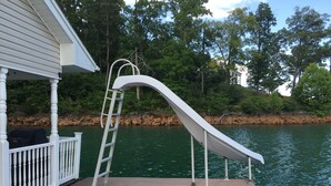 A wooden deck with a white metal slide leading into a body of water, surrounded by trees and a rocky shoreline.