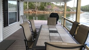 A covered deck with a table and chairs, overlooking a body of water and a wooded area.