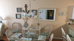 A dining area with a glass table surrounded by wicker chairs with green cushions. There are framed pictures on the wall, a chandelier, and a welcome sign.