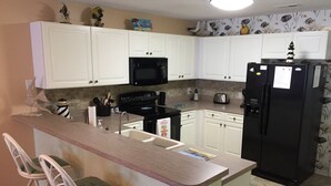 A compact kitchen with white cabinets, a black refrigerator, a microwave, and a stove. There are two bar stools with green cushions.