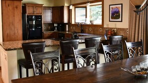 Una cocina con armarios de madera, un refrigerador negro, una zona de comedor con una mesa y sillas de madera, y un ventanal con vista a la vegetación.