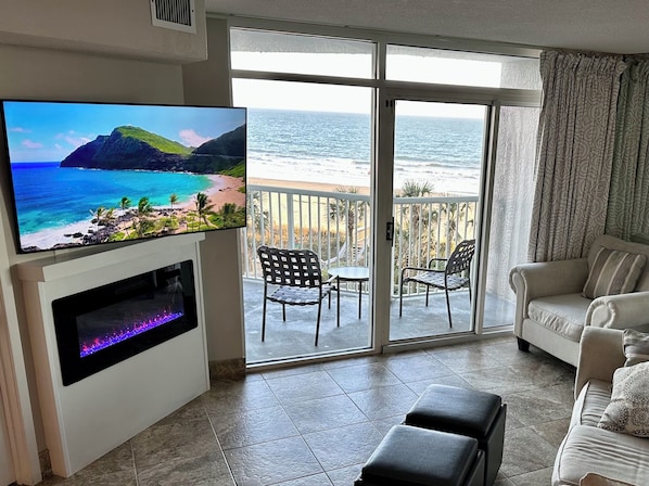 A living room with a TV, a sofa, a balcony with a view of the ocean, and a small table with chairs.