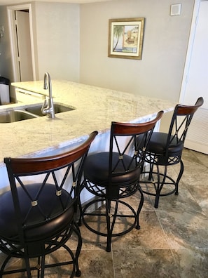 A kitchen with a granite countertop, a stainless steel sink, and three black bar stools with wooden seats.