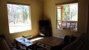 A living room with a TV, wooden furniture, and large windows with a forest view. - More than just a cabin... Experience Montana Adventure (Trout Creek)