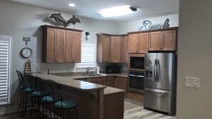 A modern kitchen with wooden cabinets, a stainless steel refrigerator, a built-in oven, and a bar area with stools. - Beautiful 6BR Gulf Front Home - "REST AREA WEST" - Pool & Deck - Reserve Now! (Gulf Shores)