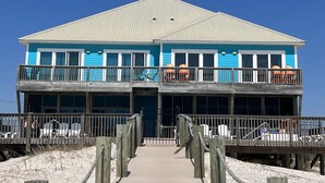A two-story building with a balcony on each floor, a wooden deck in front, and a sandy beach in the background. - Beautiful 6BR Gulf Front Home - "REST AREA WEST" - Pool & Deck - Reserve Now! (Gulf Shores)