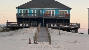 A two-story building with a balcony on the upper level, a wooden walkway leading to the entrance, and a sandy beach in the foreground. - Beautiful 6BR Gulf Front Home - "REST AREA WEST" - Pool & Deck - Reserve Now! (Gulf Shores)