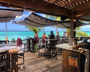 An outdoor dining area with wooden tables and chairs, a bar counter, and a view of the ocean.