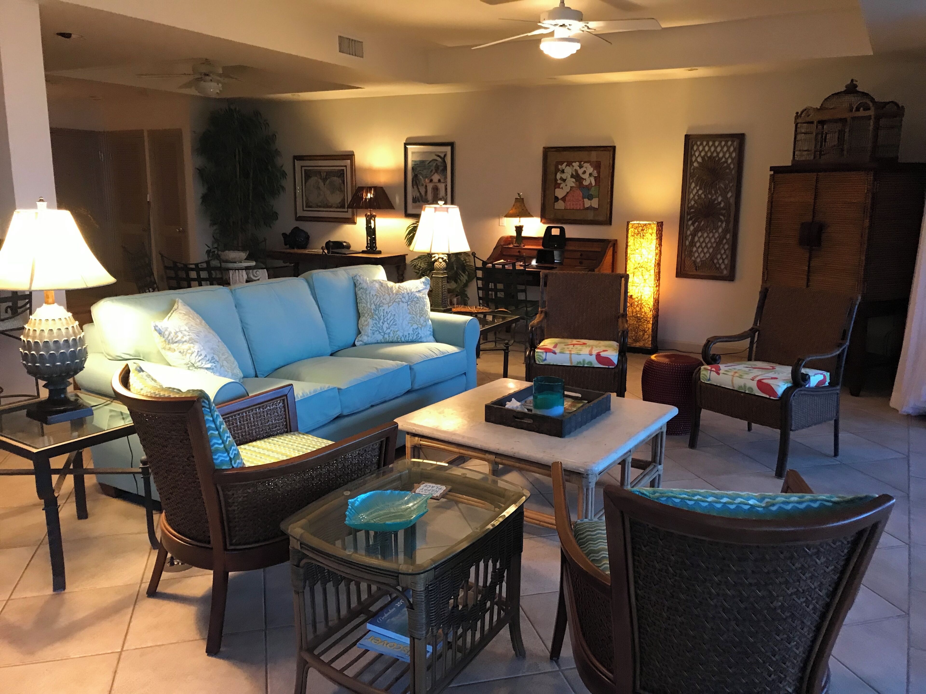 A living room with a blue sofa, wicker chairs, a coffee table, and lamps. There are framed pictures on the wall and a ceiling fan.
