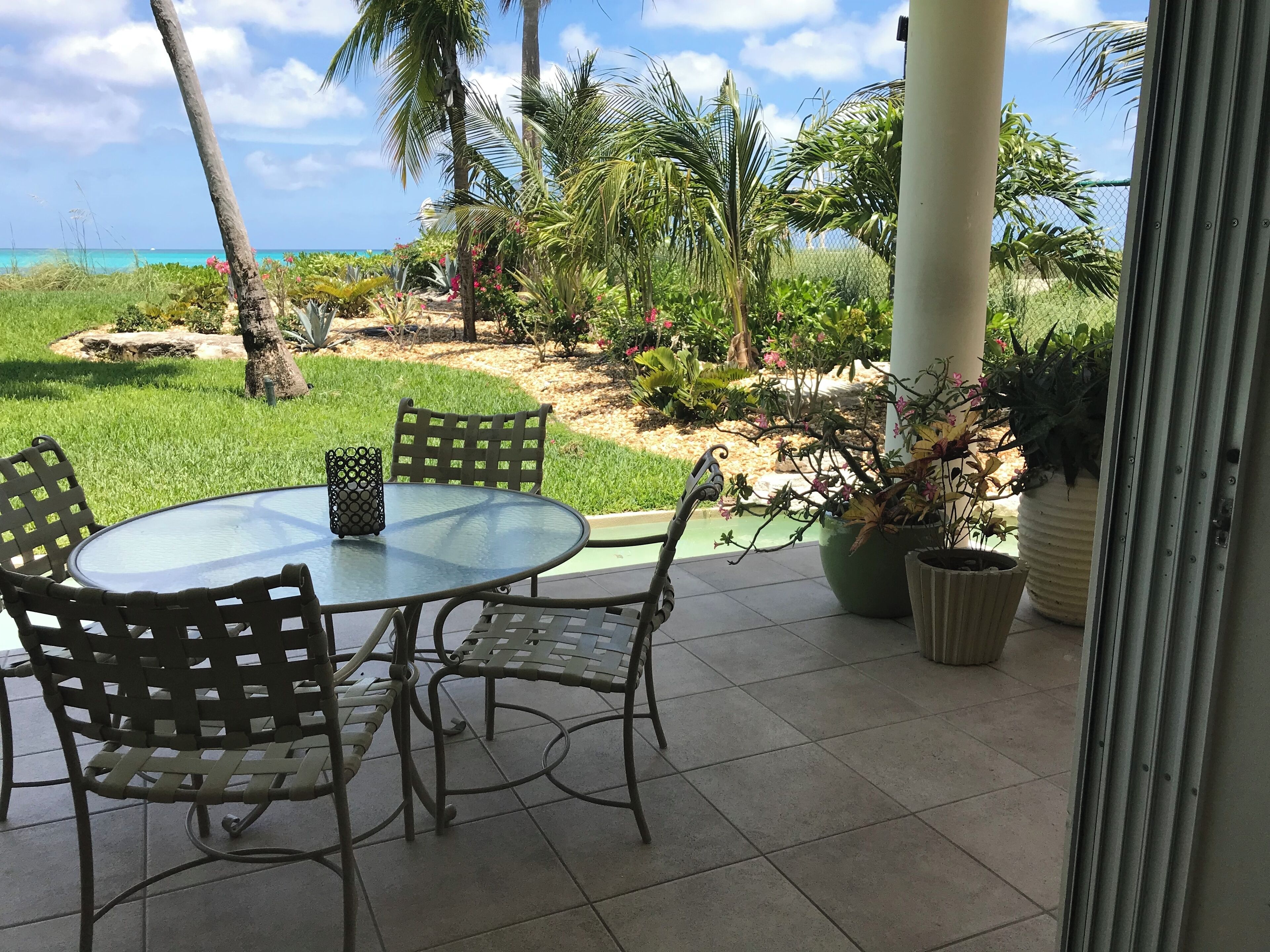 Eine Terrasse mit einem runden Glastisch und Metallstühlen, umgeben von Blumentöpfen und Blick auf das Meer.
