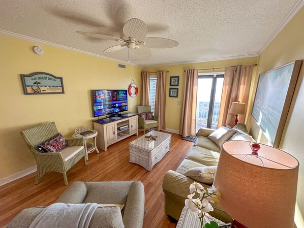 A living room with a television, wicker furniture, a ceiling fan, and a large window with curtains. - On Private Beach, Amazing View 2BR 2BA OFV Condo (Myrtle Beach)
