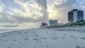 Une plage où l’on voit des empreintes de pas menant à un immeuble à étages, doté de balcons.