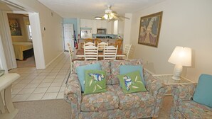 A living room with a patterned sofa set, a dining table with chairs, a kitchen area, and a ceiling fan. - Hatteras Heaven® - Oceanfront 2BR/2 Bath Condo - Pool, Renovated Kitchen, King  (Rodanthe)