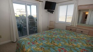 A bedroom with a bed, a television mounted on the wall, a dresser, and a sliding glass door leading to an outdoor area with chairs. - Hatteras Heaven® - Oceanfront 2BR/2 Bath Condo - Pool, Renovated Kitchen, King  (Rodanthe)