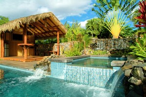Une piscine avec une cascade, une structure à toit de chaume et des plantes tropicales.