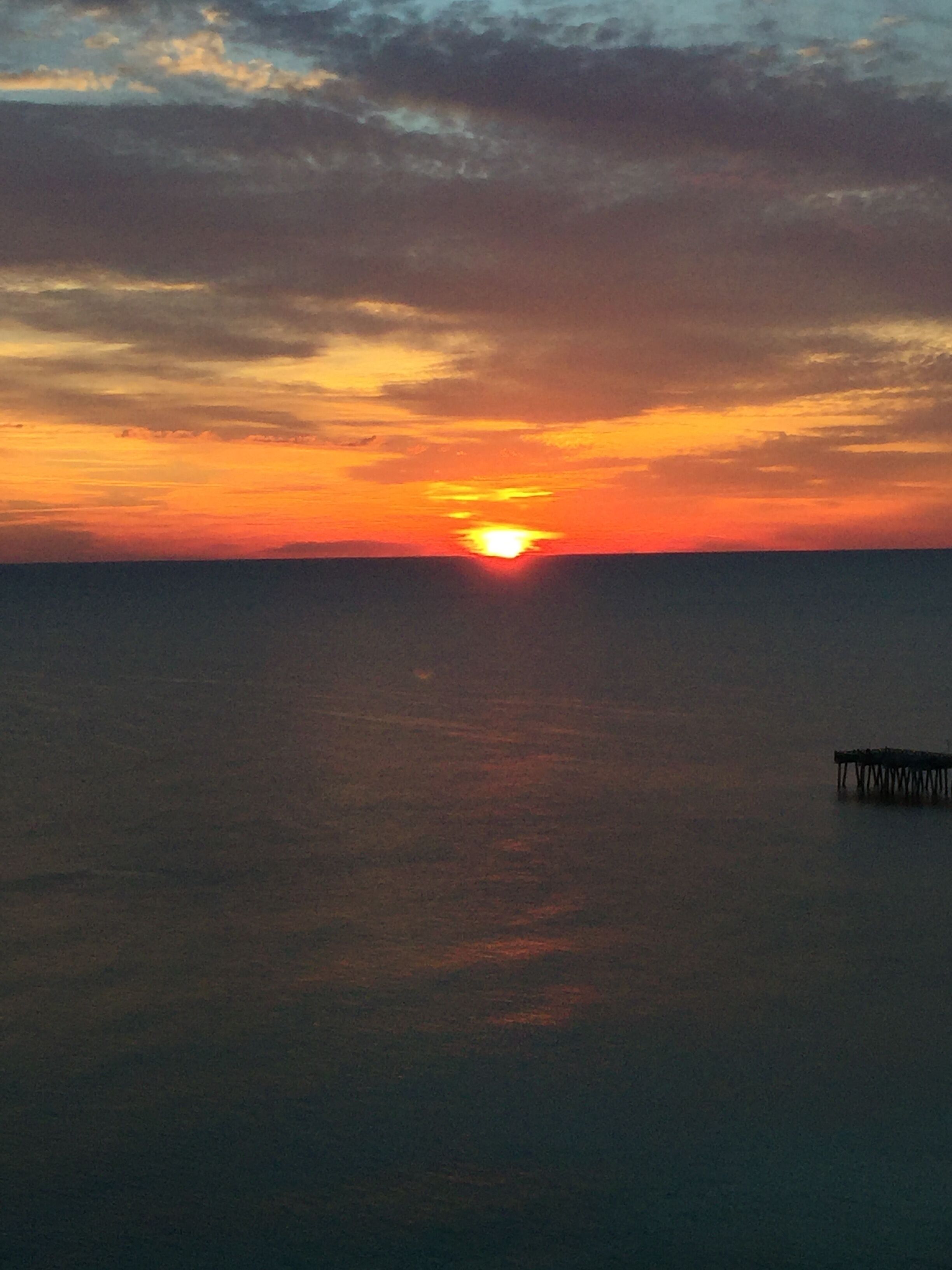 Un coucher de soleil sur une mer calme, avec une jetée qui s’avance dans l’eau.