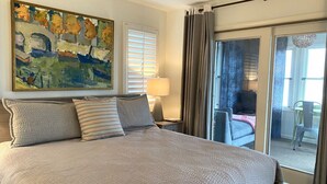 A large bed with a textured blanket, a bedside table with a lamp, a painting on the wall, and a view through a glass door to an outdoor area. - 476 Tarpon - Ocean Front - Full Time Beach (Fripp Island)