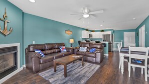 A living room with a brown leather sofa, a coffee table, and a fireplace. There is a kitchen area with white cabinets and a dining table with white chairs. -  GROUND LEVEL ON MAIN CHANNEL- WATER FRT- 4 BDRM/3 BA SLEEPS 12 WIFI (Osage Beach)