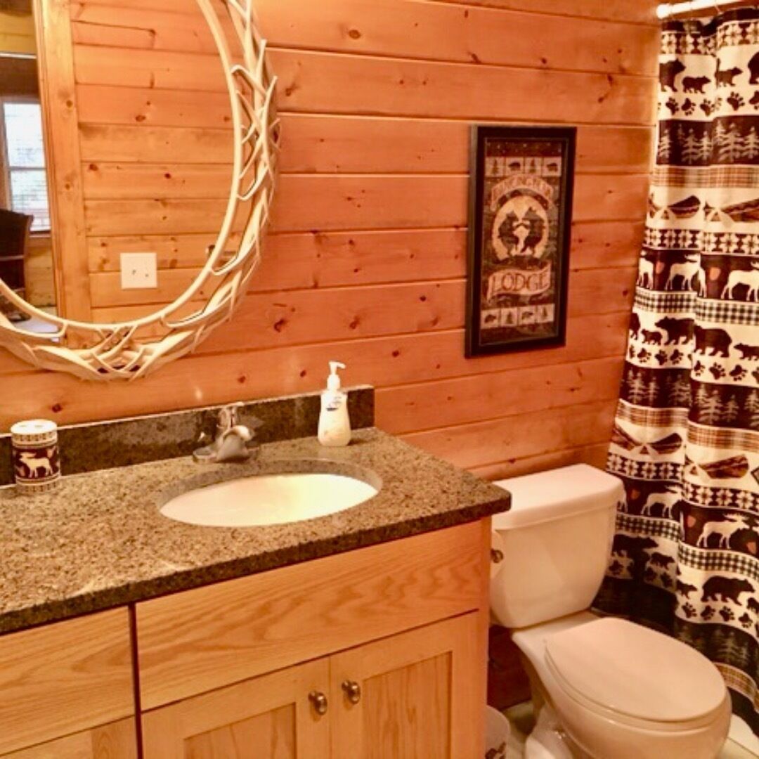 Un baño de cabaña de madera con lavamanos de granito, un cuadro en la pared y cortina de ducha estampada.