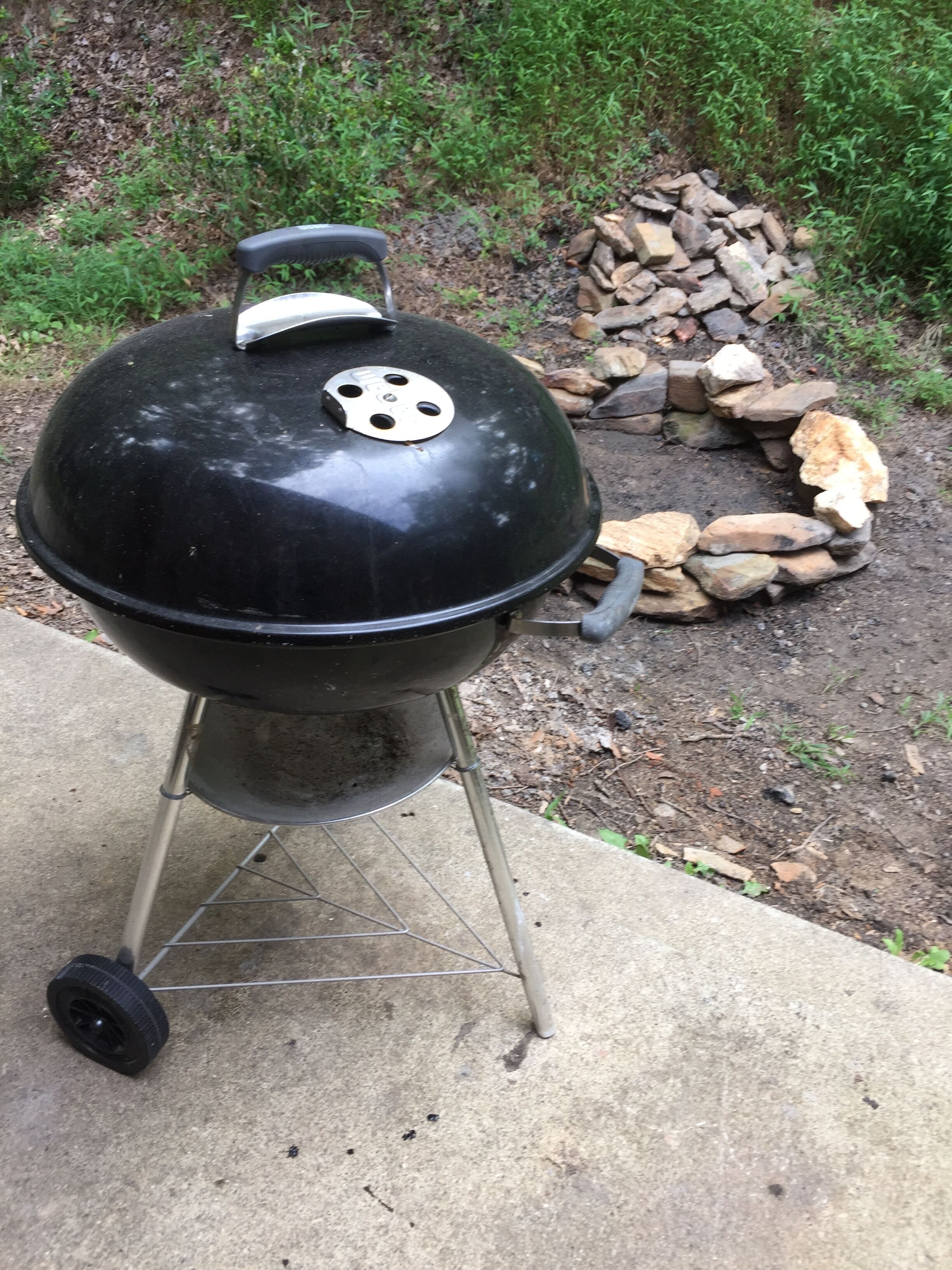 Ein schwarzer Grill mit Deckel, einem Tragegriff und einem Lüftungsschlitz obenauf. Er steht auf einer Betonfläche, im Hintergrund ist ein Holzstoß zu sehen.