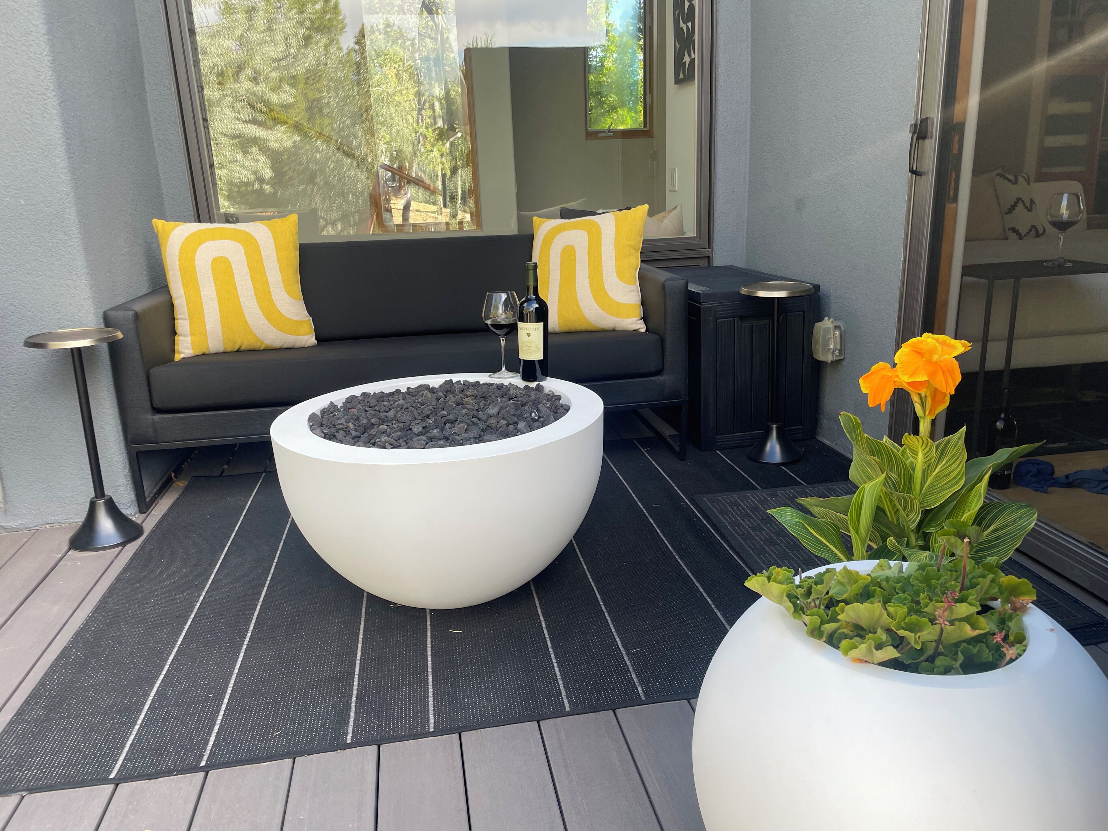 Een moderne zithoek buiten met een ronde tafel, een bank met gele kussens en een bloempot met plant.