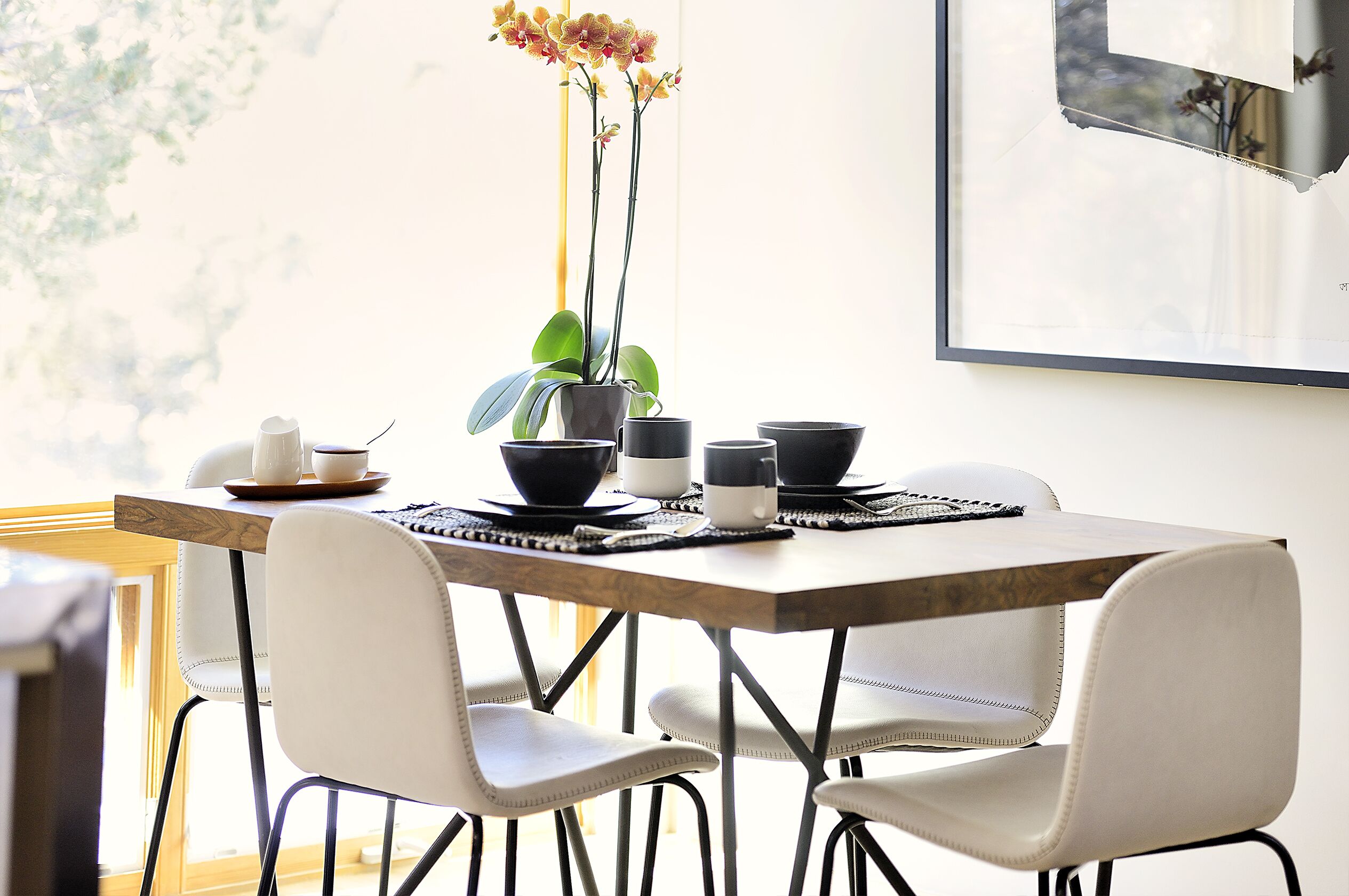 A dining area with a wooden table, white chairs, black and white cups, and a potted orchid.