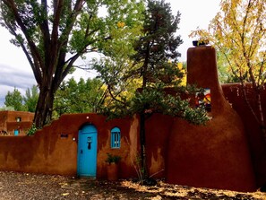 A rustic building with a blue door and arched windows, surrounded by trees and a large tree in the foreground.