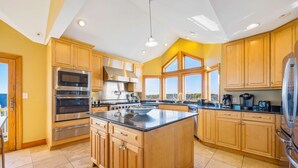 A modern kitchen with wooden cabinets, stainless steel appliances, a large island, and large windows offering a view. - Premier Oceanfront Home with Pool, Hot Tub, Elevator, Game Room, and Theater (Waves)