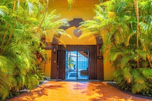 Une entrée aux allures tropicales, avec un chemin en briques menant à une porte en verre, le tout entouré d’une végétation luxuriante.