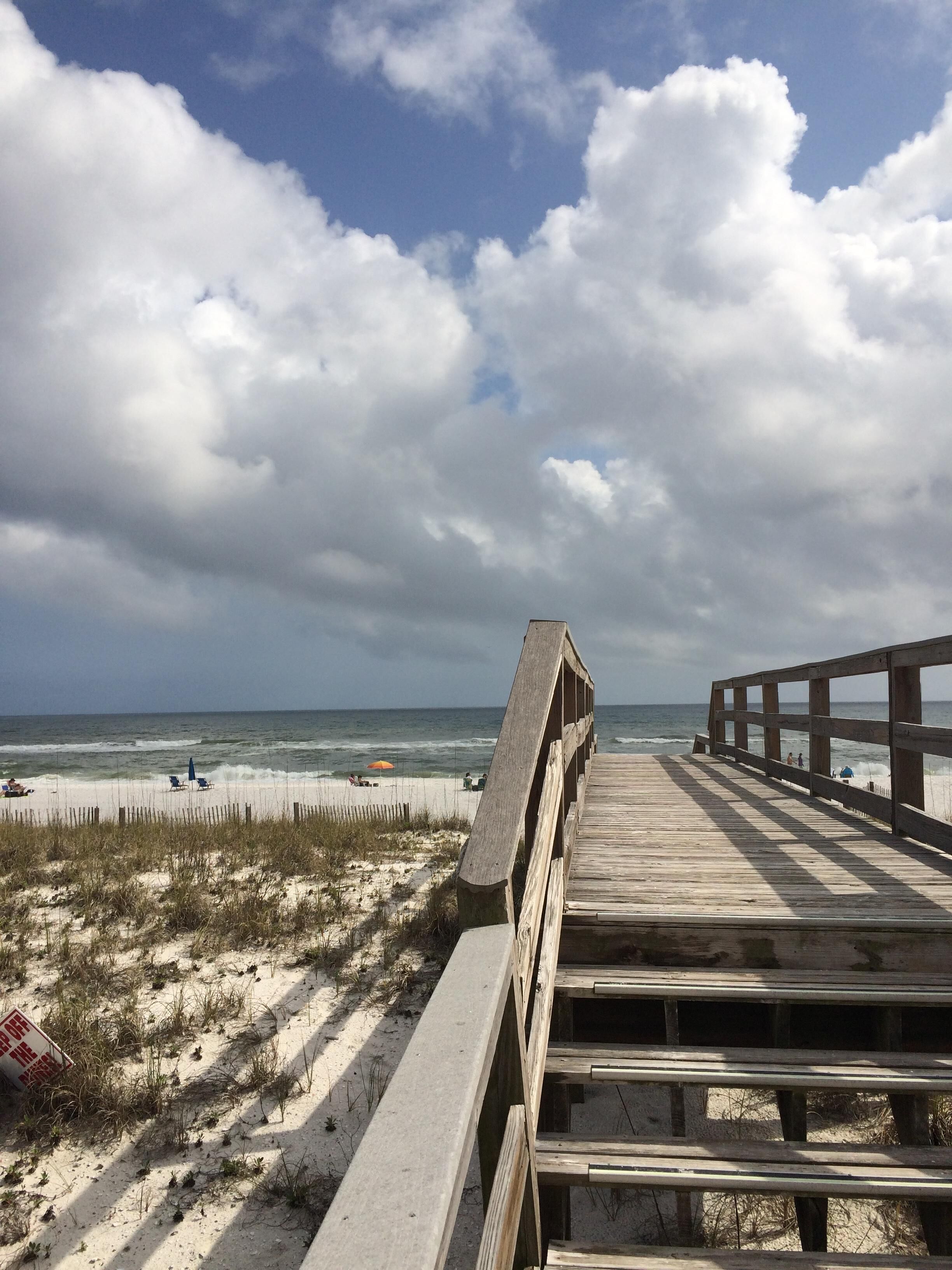 Une promenade en bois mène à une plage dont le premier plan est constitué de sable et le ciel est nuageux.