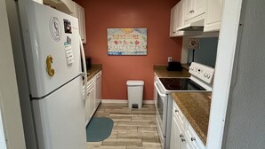 Eine kompakte Küche mit weißen Schränken, einem Kühlschrank, einem Backofen und einer Mikrowelle. Auf dem Boden liegt ein kleiner Teppich, und ein Wandbild ist sichtbar.
